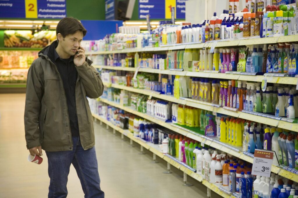 Still aus dem Dokumentarfilm "Recipes for Disaster". Mann telefoniert mit Handy im Supermarkt. Im Hintergrund ein Regal mit Reinigunsmitteln in Plastikflaschen.