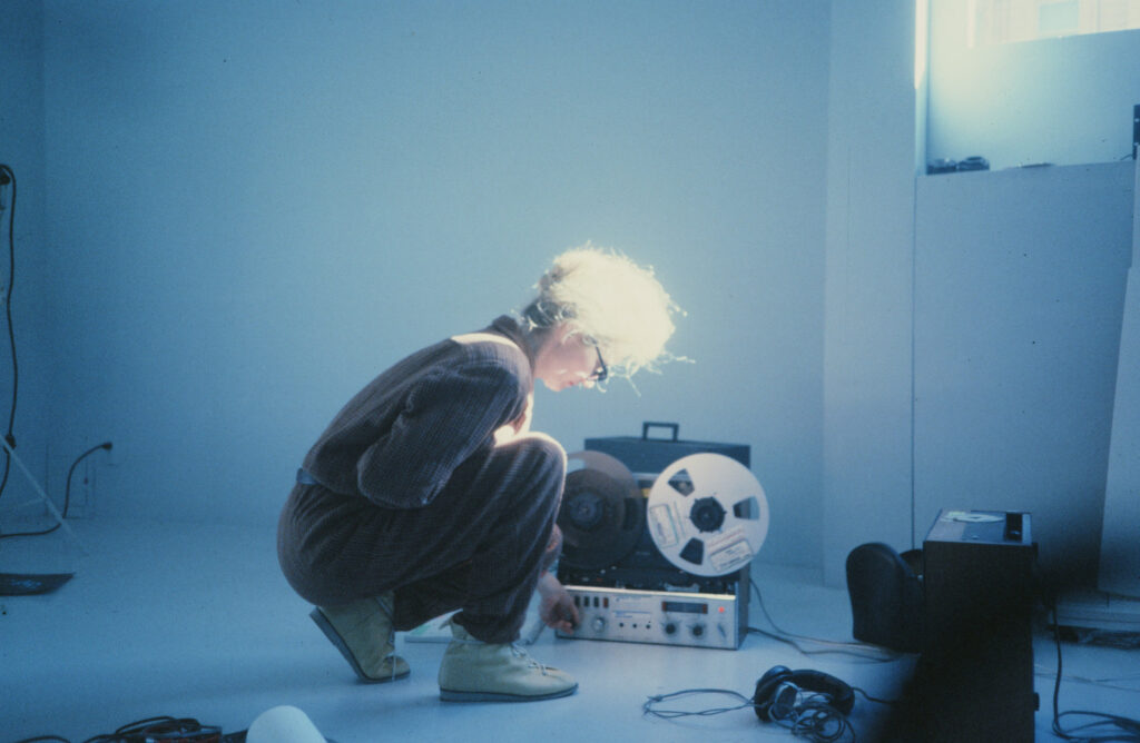 Still aus dem Dokumentarfilm "Sisters With Transistors". Eine Frau, Maryanne Amacher, kniet neben einem Tonbandgerät und bedient dieses.