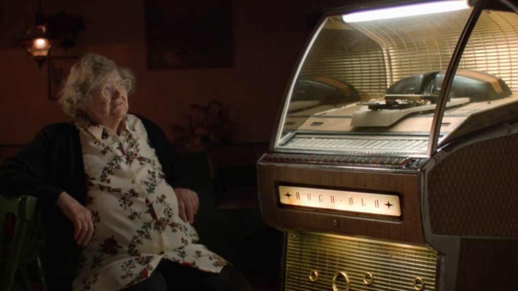Szene aus dem Dokumentarfilm 80.000 Schnitzel. Eine alte Frau sitzt neben einer Musicbox und hört Musik aus den 1960er Jahren.