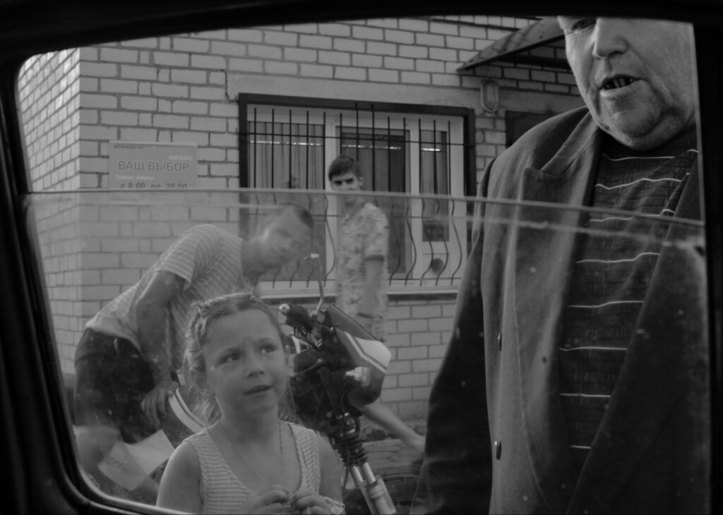 Still aus dem Dokumentarfilms KRAI. Ein kleines Mädchen und ein alter Mann schauen zum Fenster eines PKW hinein.