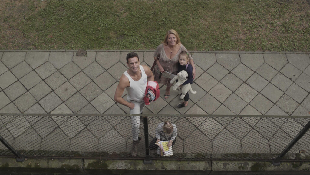 Still aus dem Dokumentarfilm THE BALCONY MOVIE. Eine Familie steht auf dem Bürgersteig und schaut zur Kamera, die auf einem Balkon im ersten Stock aufgestellt ist.