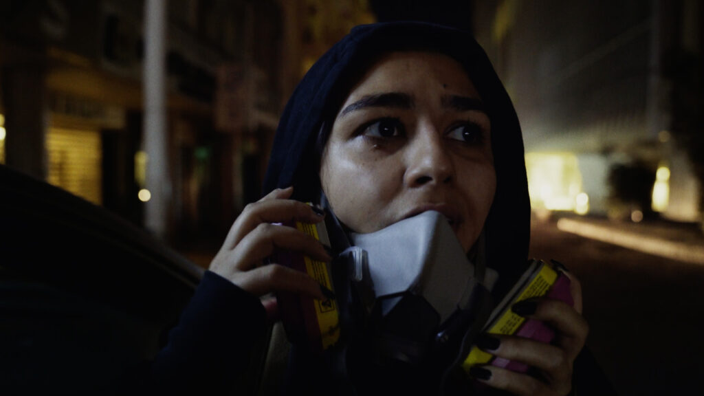 Still aus dem Dokumentarfilm ANXIOUS IN BEIRUT. Zu sehen ist eine junge Frau mit Kapuzenpullover und Gasmaske am Rande einer Straßenschlacht in Beirut.