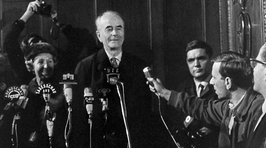 Still aus dem Dokumentarfilm "Speer goes to Hollywood". Albert Speer steht vor zahlreichen Mikrofonen. Er gibt ein Interview.