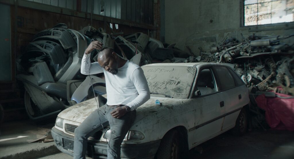 Still aus dem Dokumentarfilms Bewegungen eines nahen Bergs von Sebastian Brameshuber. Ein mann sitzt in einer Lagerhalle auf der Motorhaube eines stark eingestaubten Autos. Er rasiert sich die Haare mit einer Haarschneidemaschine.