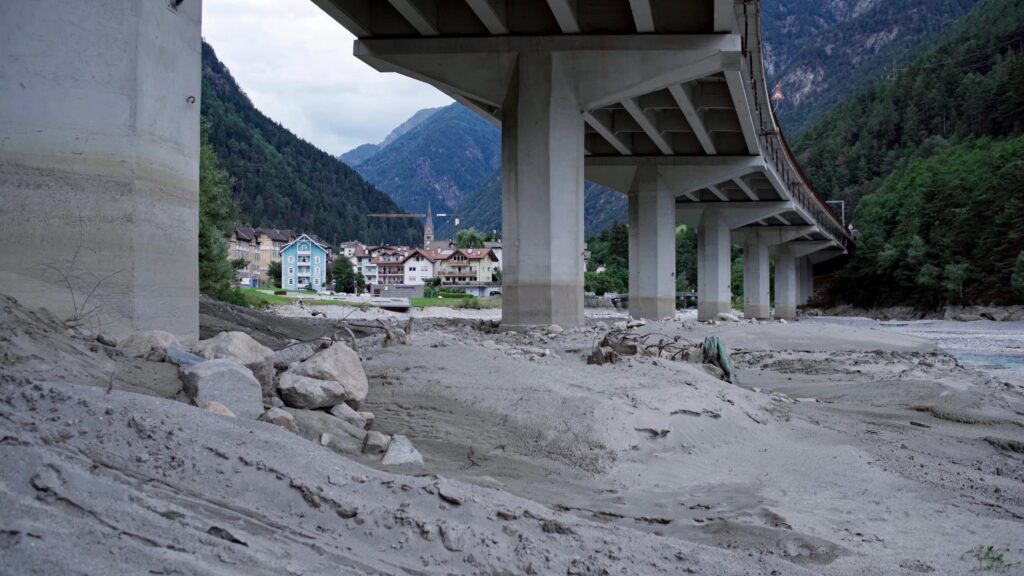 Still aus dem Dokumentarfilm Durchgangsland von Daniel Fill. Im Vordergrund ist eine Autobahnbrücke zu sehen die einen Bergfluss überquert. Im Hintergrund sieht man eine Ortschaft (Franzenfeste) und die Alpen.