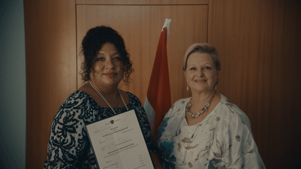 Still aus dem Dokumentarfilm "Noch lange keine Lippizaner" von Olga Kosanović. Eine Sachbearbeiterin überreicht einer Frau die Urkunde über die Staatsbürgerschaft in Österreich.