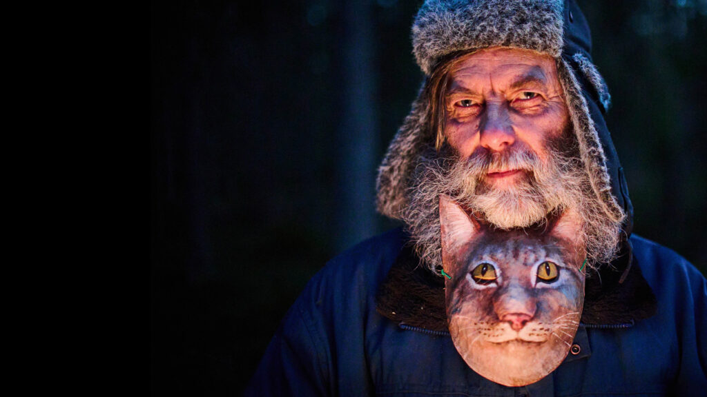 Still aus dem Dokumentarfilm "Lynx Man". Der Finne Hannu posiert mit einer Luchs-Maske, die um seinen Hals hängt, für die Kamera.
